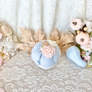 May include: A light blue fabric pumpkin with a pink flower accent sits on a white lace tablecloth. The pumpkin is surrounded by fall-themed decorations, including dried leaves, a small blue gourd, and pink and white flowers.