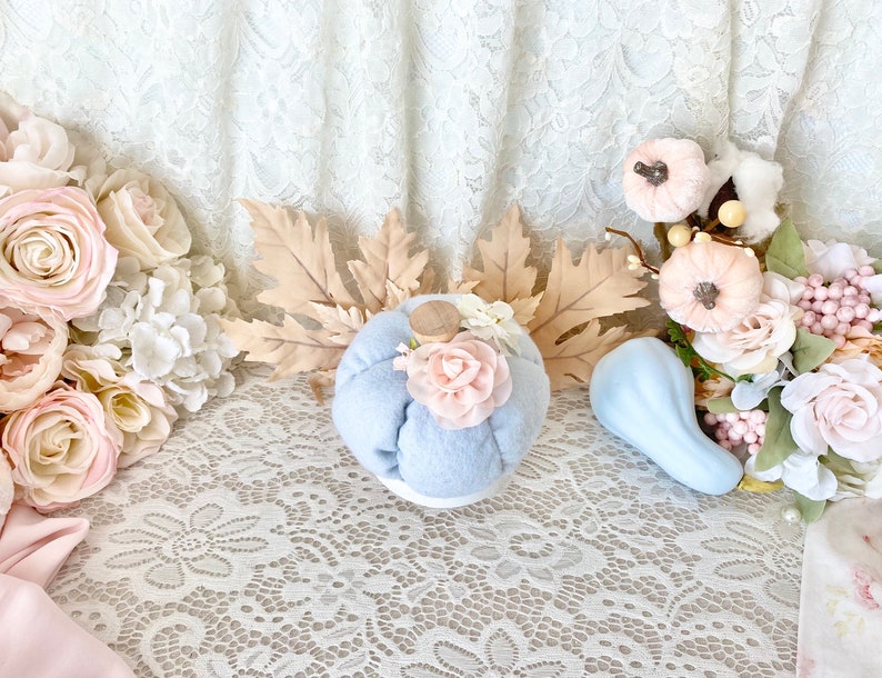 May include: A light blue fabric pumpkin with a pink flower accent sits on a white lace tablecloth. The pumpkin is surrounded by fall-themed decorations, including dried leaves, a small blue gourd, and pink and white flowers.