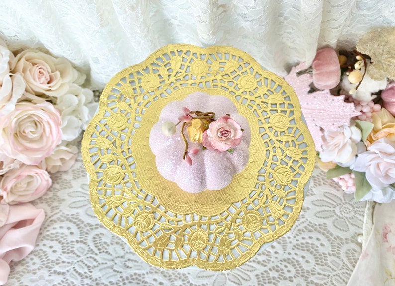 May include: A pink glittery pumpkin with a floral embellishment sits on a gold paper doily.