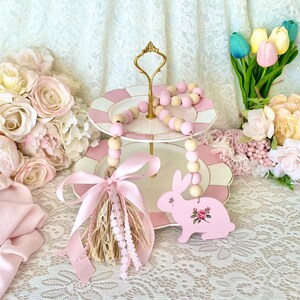 Pu&ograve; includere: Un vassoio a due livelli con piatti rosa e bianchi, adornato con una fila di perline di legno rosa e naturali. Un ornamento a forma di coniglietto rosa con dettagli floreali &egrave; appeso sotto. La scena &egrave; decorata con rose e tulipani rosa.