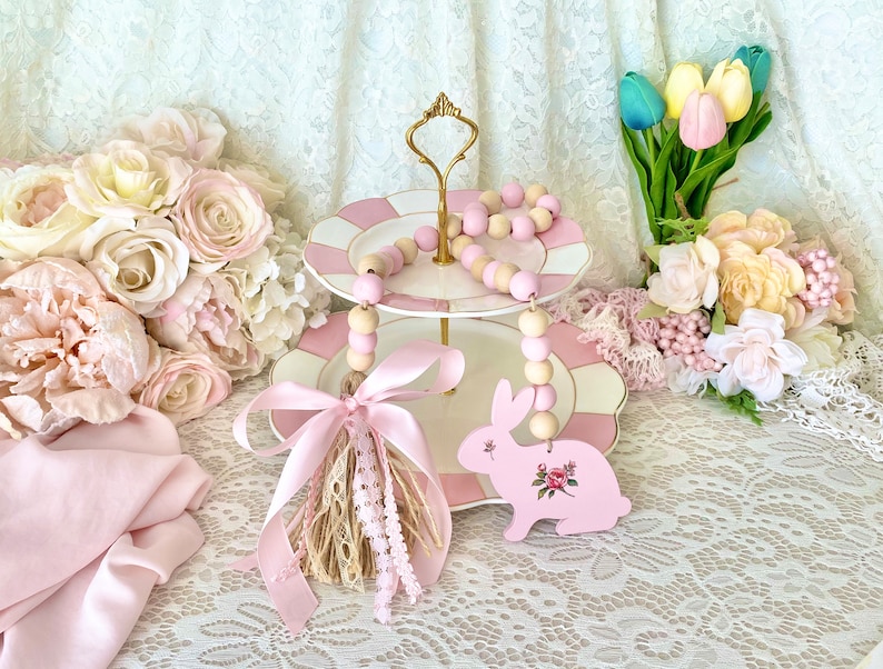 Pu&ograve; includere: Un vassoio a due livelli con piatti rosa e bianchi, adornato con una fila di perline di legno rosa e naturali. Un ornamento a forma di coniglietto rosa con dettagli floreali &egrave; appeso sotto. La scena &egrave; decorata con rose e tulipani rosa.