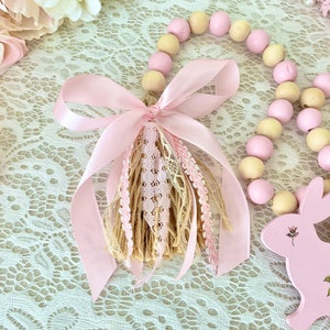 Pu&ograve; includere: Una ghirlanda decorativa con perline di legno rosa e crema alternate. Un nastro rosa e un nappa con dettagli in pizzo e iuta adornano la ghirlanda. &Egrave; presente anche un coniglietto di legno rosa con dettagli floreali.