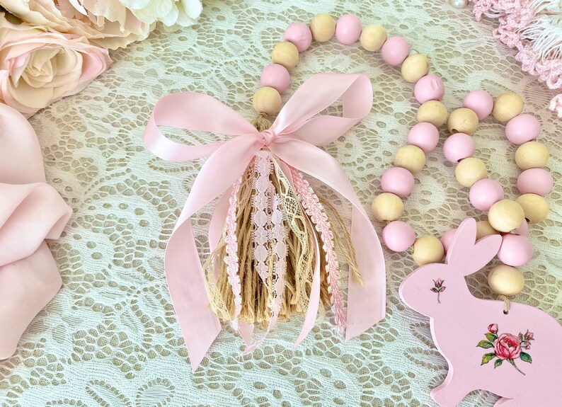 Pu&ograve; includere: Una ghirlanda decorativa con perline di legno rosa e crema alternate. Un nastro rosa e un nappa con dettagli in pizzo e iuta adornano la ghirlanda. &Egrave; presente anche un coniglietto di legno rosa con dettagli floreali.
