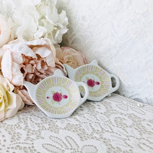 May include: Two white ceramic tea bag holders shaped like teapots with floral designs. The holders are on a white lace tablecloth.