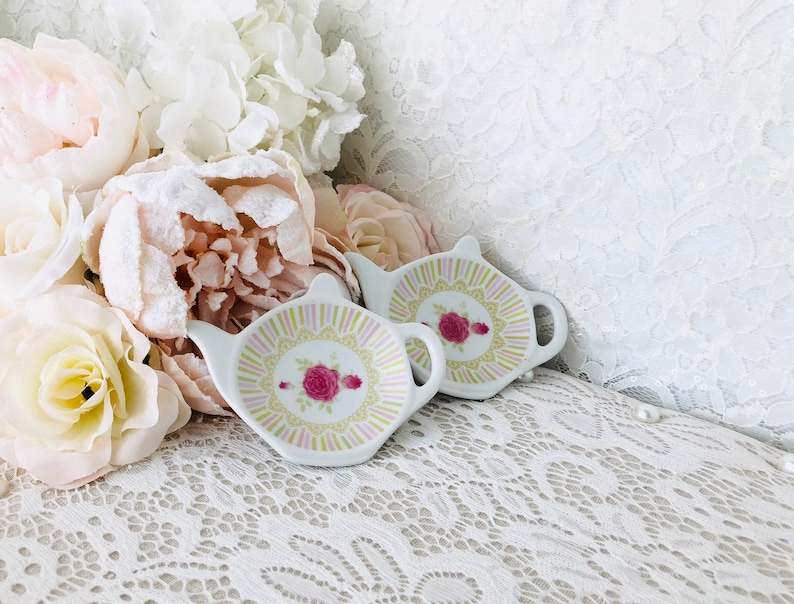 May include: Two white ceramic tea bag holders shaped like teapots with floral designs. The holders are on a white lace tablecloth.