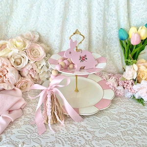 Pu&ograve; includere: Un alzatina a due livelli con un design a righe rosa e bianche. Un ornamento a forma di coniglio rosa con dettagli floreali &egrave; posto sopra. L'alzatina ha una maniglia dorata ed &egrave; circondata da fiori rosa e un nappa.