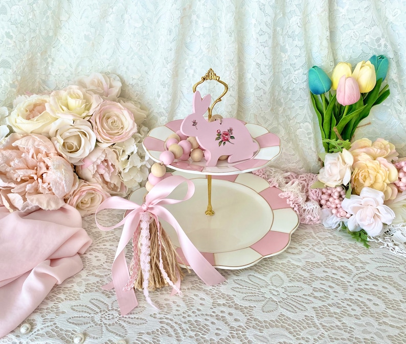 Pu&ograve; includere: Un alzatina a due livelli con un design a righe rosa e bianche. Un ornamento a forma di coniglio rosa con dettagli floreali &egrave; posto sopra. L'alzatina ha una maniglia dorata ed &egrave; circondata da fiori rosa e un nappa.