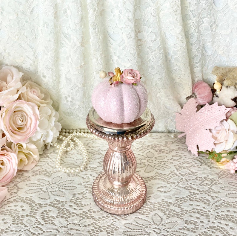 May include: A pink glitter pumpkin with a floral accent sits on a rose gold pedestal. The pumpkin is on a white lace tablecloth.