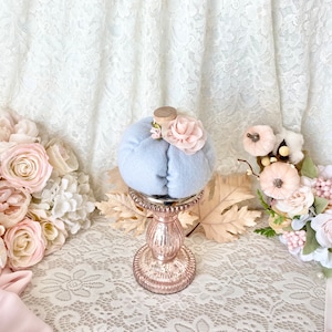 May include: A light blue fabric pumpkin with a pink flower on top, sitting on a gold metal stand with a decorative base. The pumpkin is surrounded by pink and white flowers.