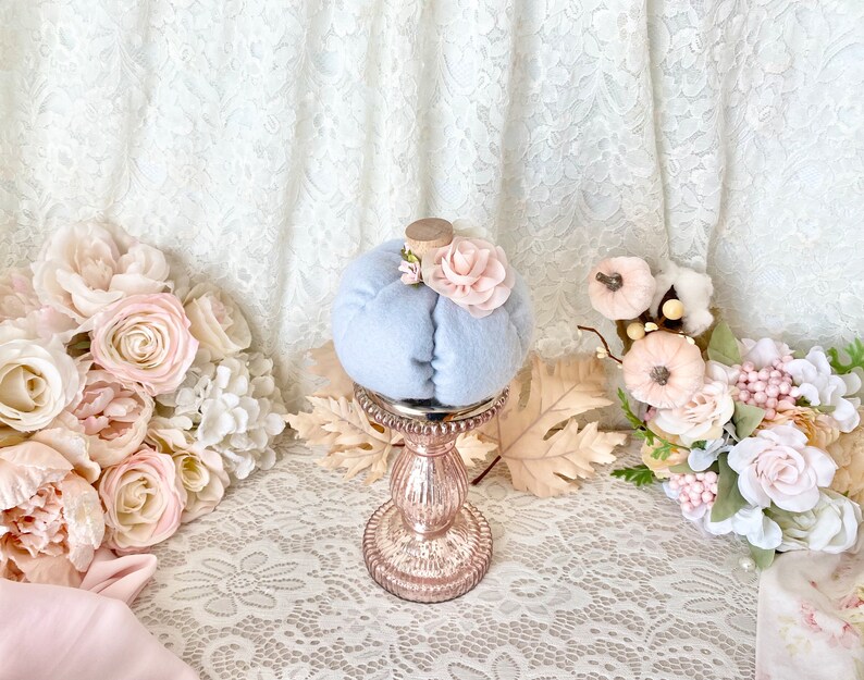 May include: A light blue fabric pumpkin with a pink flower on top, sitting on a gold metal stand with a decorative base. The pumpkin is surrounded by pink and white flowers.