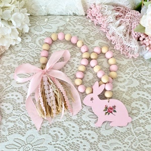 Pu&ograve; includere: Una ghirlanda decorativa di perline di legno con un ciondolo a forma di coniglietto rosa. La ghirlanda presenta perline rosa e crema alternate. Il ciondolo a forma di coniglietto &egrave; dipinto di rosa e decorato con un motivo floreale. Un nastro rosa e un nappa aggiungono un tocco decorativo.