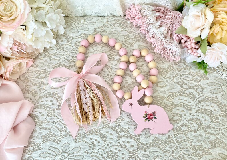 Pu&ograve; includere: Una ghirlanda decorativa di perline di legno con un ciondolo a forma di coniglietto rosa. La ghirlanda presenta perline rosa e crema alternate. Il ciondolo a forma di coniglietto &egrave; dipinto di rosa e decorato con un motivo floreale. Un nastro rosa e un nappa aggiungono un tocco decorativo.