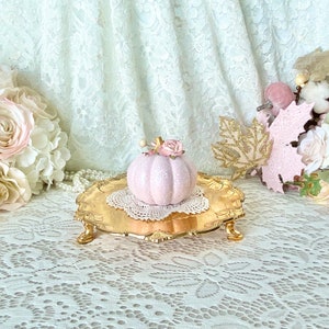 May include: A pink ceramic pumpkin with a floral accent sits on a gold decorative stand. The stand is on a white lace tablecloth.