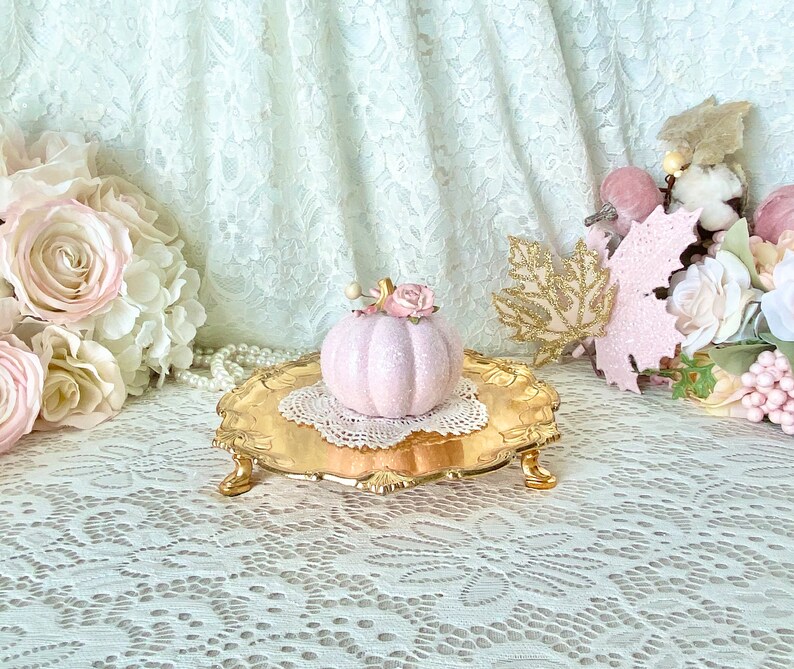 May include: A pink ceramic pumpkin with a floral accent sits on a gold decorative stand. The stand is on a white lace tablecloth.