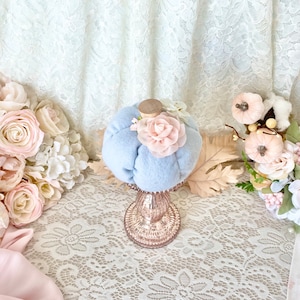 May include: A light blue fabric pumpkin with a pink fabric flower on top, sitting on a glass pedestal with a lace tablecloth underneath. The pumpkin is surrounded by pink and white flowers.