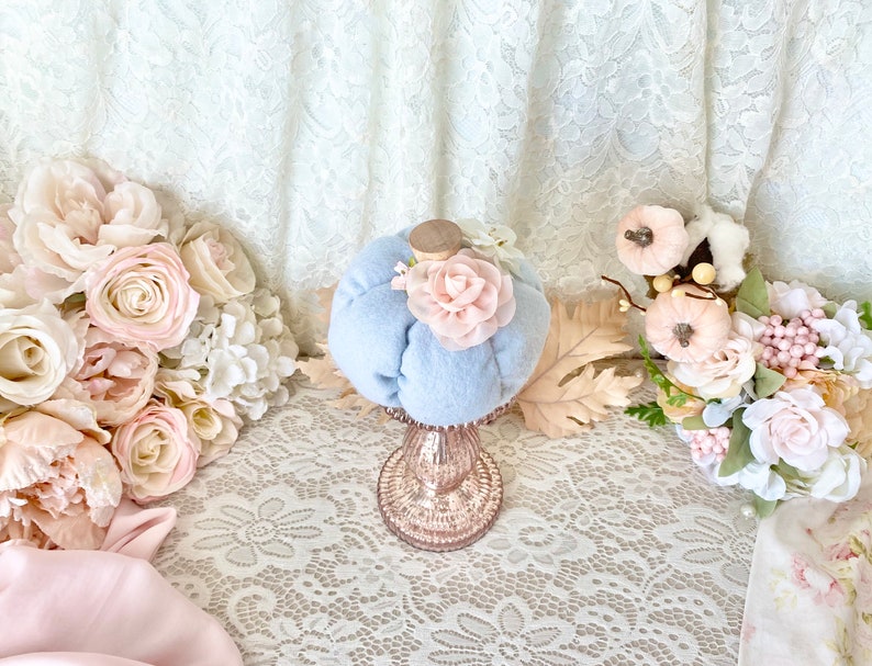 May include: A light blue fabric pumpkin with a pink fabric flower on top, sitting on a glass pedestal with a lace tablecloth underneath. The pumpkin is surrounded by pink and white flowers.