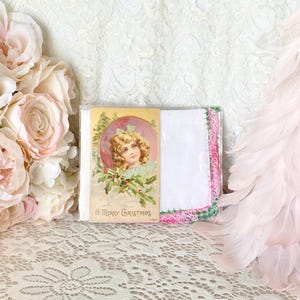 May include: A vintage Christmas card with a young girl in a pink dress and a green wreath. The card says "A Merry Christmas". The card is next to a white handkerchief with pink and green lace trim.