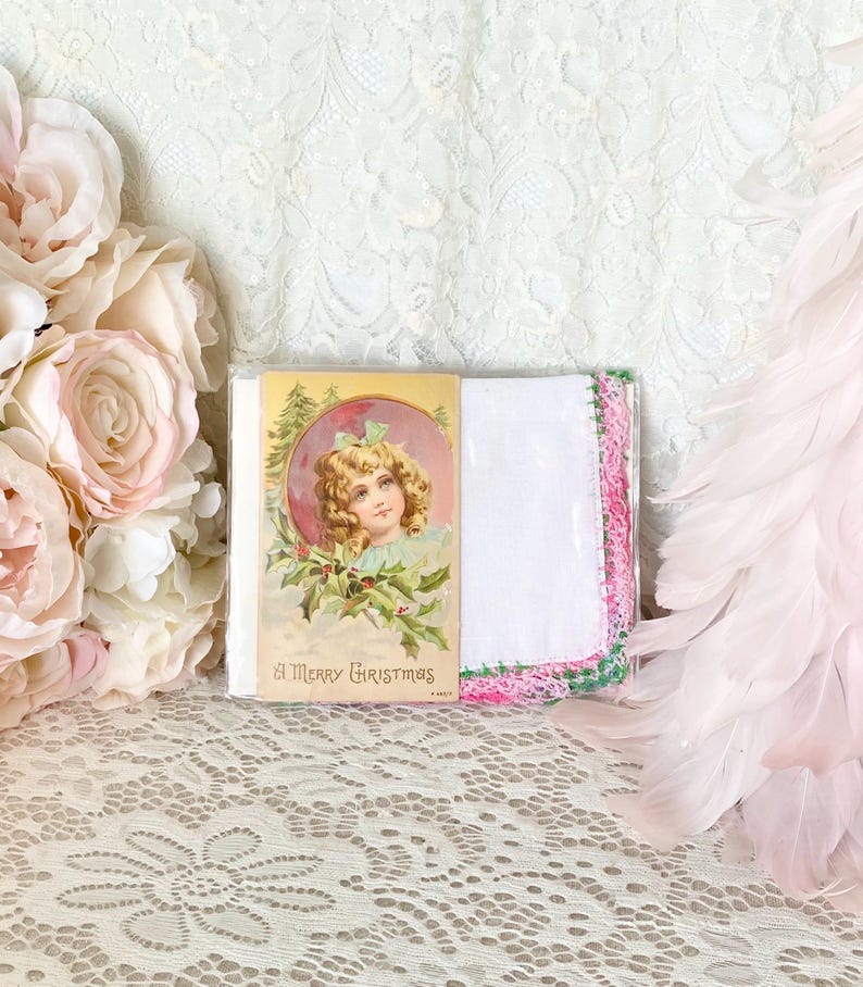 May include: A vintage Christmas card with a young girl in a pink dress and a green wreath. The card says "A Merry Christmas". The card is next to a white handkerchief with pink and green lace trim.