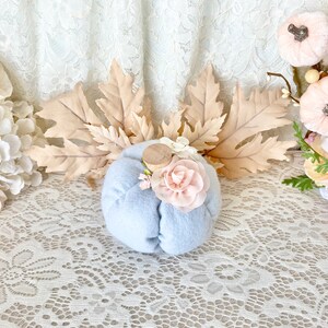 May include: A light blue felt pumpkin with a wooden stem and pink fabric flowers. The pumpkin is sitting on a white lace tablecloth with fall leaves and other decorative pumpkins in the background.