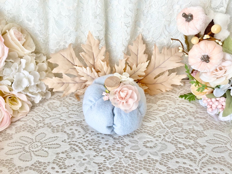 May include: A light blue felt pumpkin with a wooden stem and pink fabric flowers. The pumpkin is sitting on a white lace tablecloth with fall leaves and other decorative pumpkins in the background.