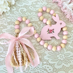 Pu&ograve; includere: Due ghirlande pasquali decorative. Una presenta un coniglietto rosa con dettagli floreali, l'altra un nappa con nastro e pizzo rosa. Entrambe sono decorate con perline di legno rosa e naturali.