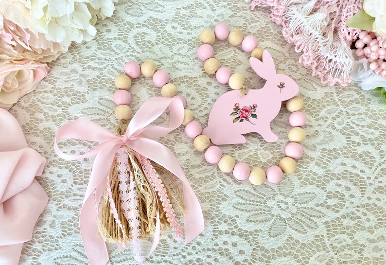 Pu&ograve; includere: Due ghirlande pasquali decorative. Una presenta un coniglietto rosa con dettagli floreali, l'altra un nappa con nastro e pizzo rosa. Entrambe sono decorate con perline di legno rosa e naturali.