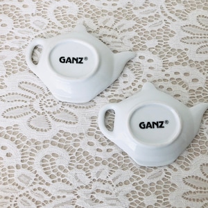 May include: Two white ceramic teapot-shaped dishes with the word "GANZ&reg;" printed on the bottom.