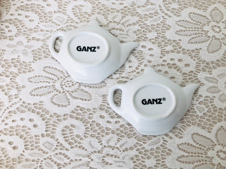 May include: Two white ceramic teapot-shaped dishes with the word "GANZ&reg;" printed on the bottom.