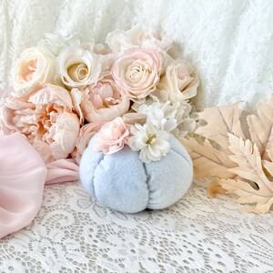 May include: A light blue felt pumpkin with white and pink flower accents. The pumpkin is sitting on a white lace tablecloth with pink and white flowers in the background.