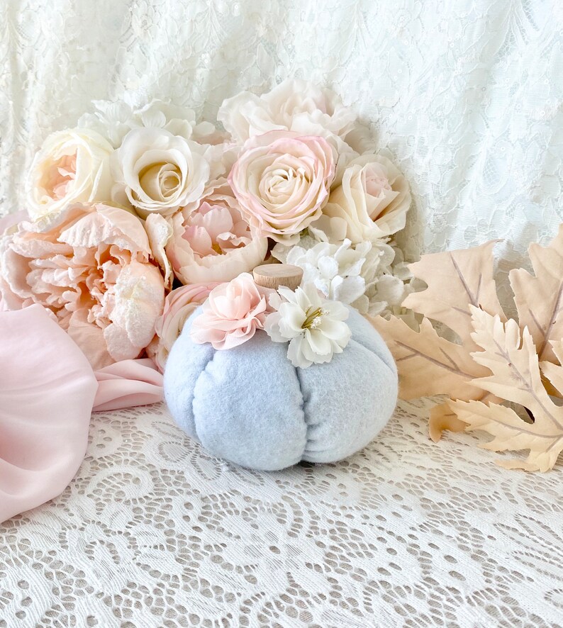 May include: A light blue felt pumpkin with white and pink flower accents. The pumpkin is sitting on a white lace tablecloth with pink and white flowers in the background.