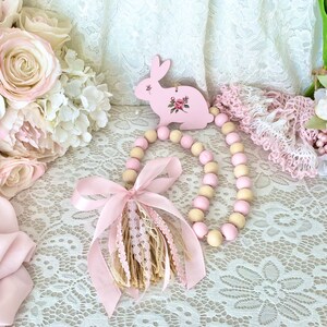 Pu&ograve; includere: Un ornamento a forma di coniglio in legno rosa con un design floreale &egrave; appeso sopra una ghirlanda di perline di legno rosa e crema. Un nastro rosa e un nappa aggiungono dettagli. La scena &egrave; ambientata su una superficie di pizzo bianco, con composizioni floreali sullo sfondo.