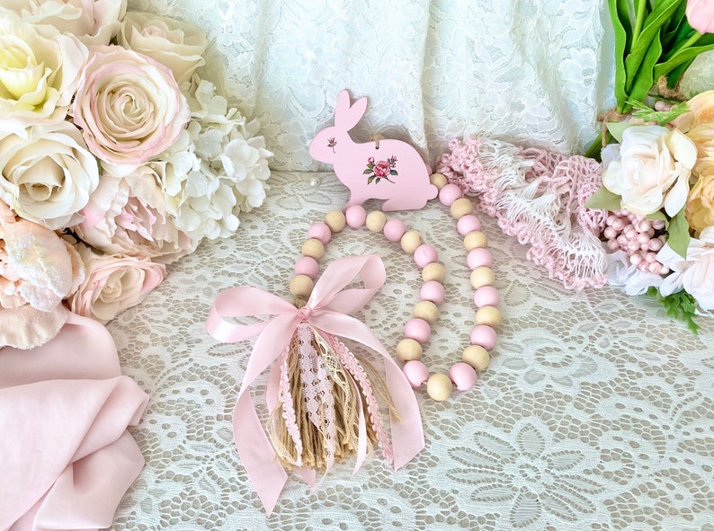 Pu&ograve; includere: Un ornamento a forma di coniglio in legno rosa con un design floreale &egrave; appeso sopra una ghirlanda di perline di legno rosa e crema. Un nastro rosa e un nappa aggiungono dettagli. La scena &egrave; ambientata su una superficie di pizzo bianco, con composizioni floreali sullo sfondo.