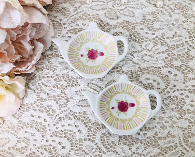 May include: Two white ceramic tea bag holders shaped like teapots. The holders have a pink, green, and yellow floral design with a pink rose in the center.