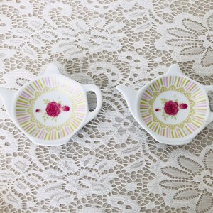 May include: Two white ceramic tea bag holders shaped like teapots with pink, green, and gold floral designs.