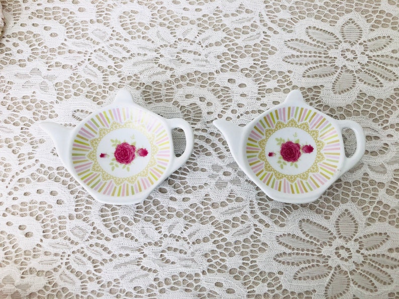 May include: Two white ceramic tea bag holders shaped like teapots with pink, green, and gold floral designs.