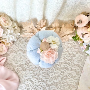 May include: A light blue felt pumpkin with a wooden top and pink and white fabric flowers. The pumpkin is sitting on a white lace tablecloth.