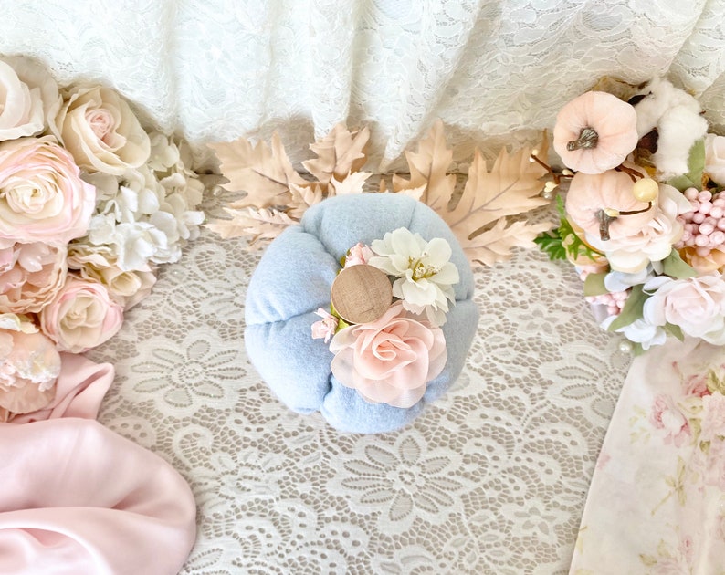 May include: A light blue felt pumpkin with a wooden top and pink and white fabric flowers. The pumpkin is sitting on a white lace tablecloth.