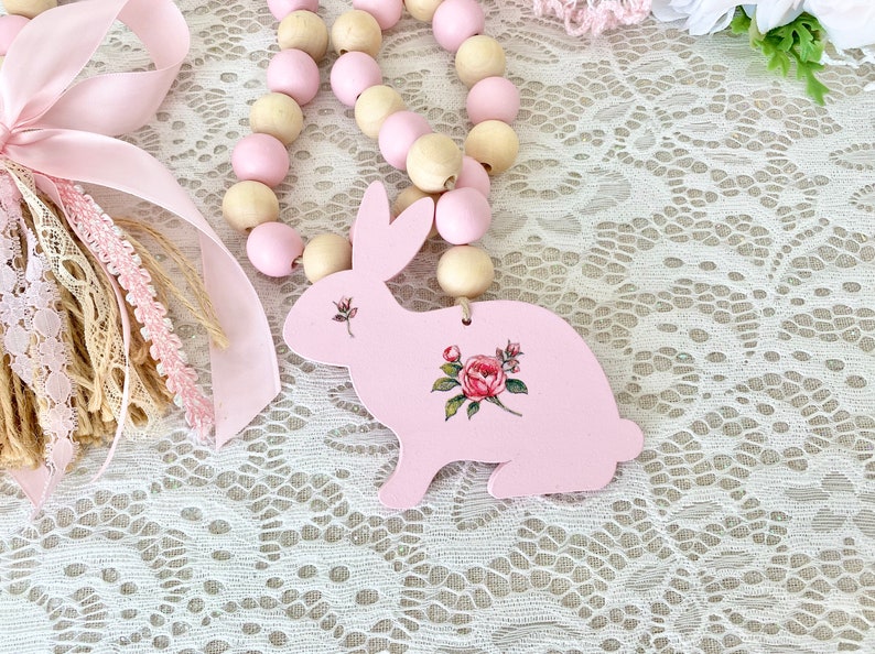 Pu&ograve; includere: Un ornamento a forma di coniglio in legno rosa con un design floreale, parte di una ghirlanda di perline. La ghirlanda presenta perline di legno rosa e naturale alternate. Sono visibili anche nastri e pizzi decorativi, che suggeriscono un tema primaverile o pasquale.