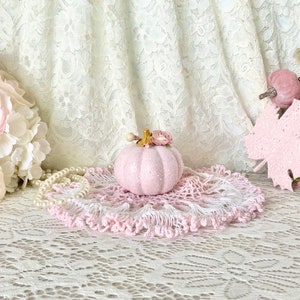 May include: A pink glitter pumpkin with a floral accent sits on a pink and white crocheted doily. The pumpkin is surrounded by white lace and pearls.