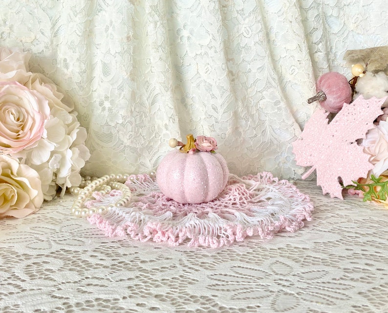 May include: A pink glitter pumpkin with a floral accent sits on a pink and white crocheted doily. The pumpkin is surrounded by white lace and pearls.