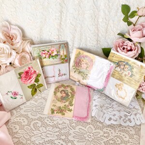 May include: A collection of vintage-style greeting cards with floral and romantic themes. The cards feature illustrations of roses, angels, and other delicate designs. The cards are displayed on a white lace tablecloth with pink roses.
