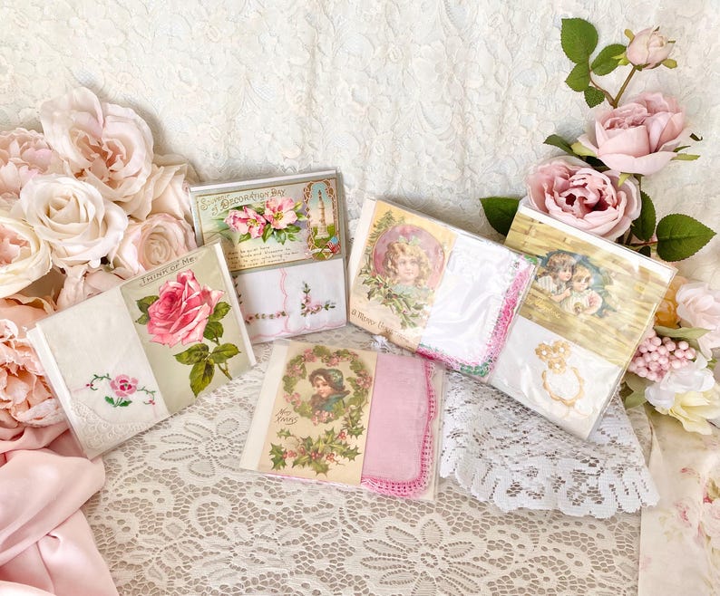 May include: A collection of vintage-style greeting cards with floral and romantic themes. The cards feature illustrations of roses, angels, and other delicate designs. The cards are displayed on a white lace tablecloth with pink roses.