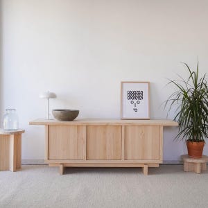 May include: A light wood cabinet with three doors and a light wood top. A white lamp with a white shade is on the left side of the cabinet. A small, round, gray bowl is on the top of the cabinet. A framed black and white print with the text "O.I.P" is on the wall behind the cabinet. A potted plant is on the right side of the cabinet.