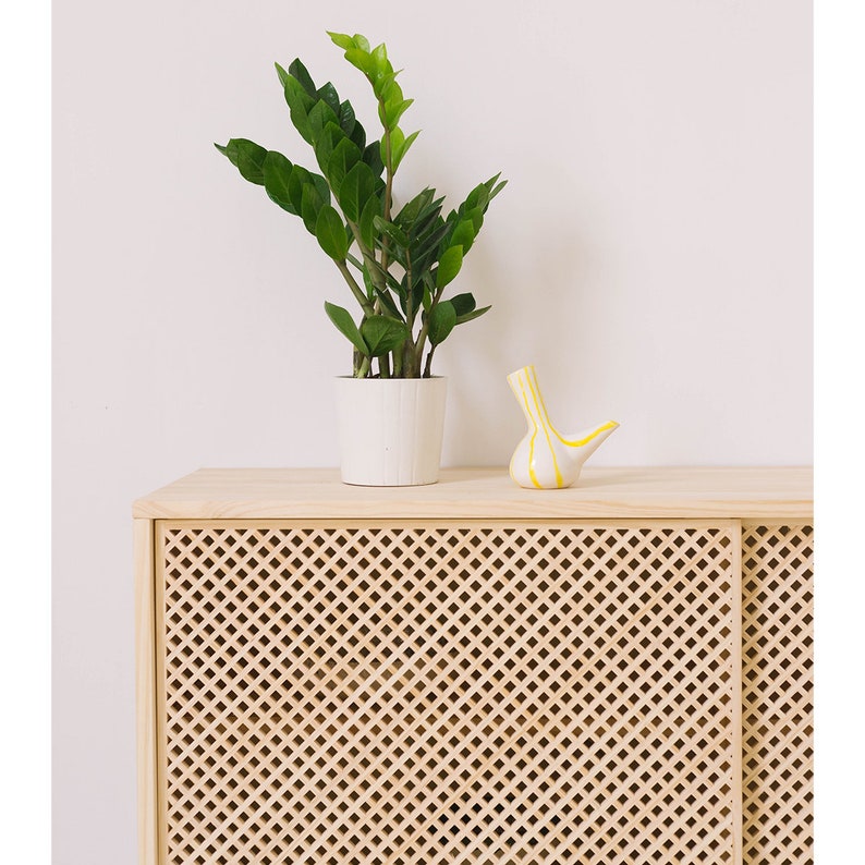 May include: A light wood cabinet with two doors featuring a diamond-shaped lattice pattern. A potted plant and a white ceramic vase with yellow stripes are on top of the cabinet.