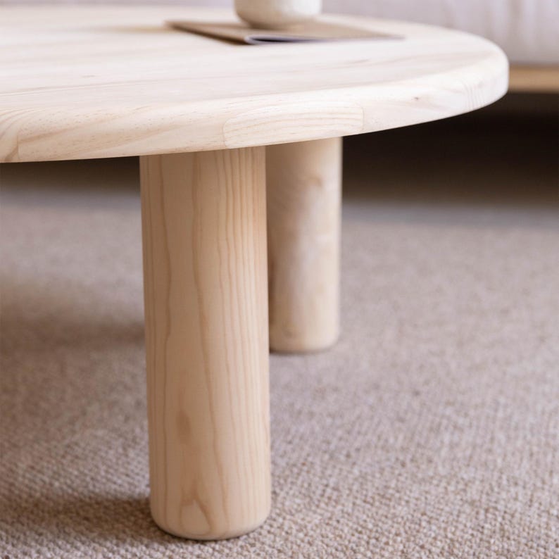 May include: A light-colored, round wooden coffee table with three cylindrical legs. The table has a smooth, natural finish and sits on a neutral-colored carpet. The table's design is minimalist and modern, suitable for a living room.