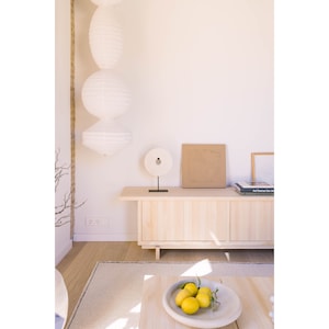 May include: A light wood cabinet with three doors and a white stone circle sculpture on top. The cabinet is against a white wall with a white paper lantern hanging from the ceiling. There are two books and a framed picture on top of the cabinet.