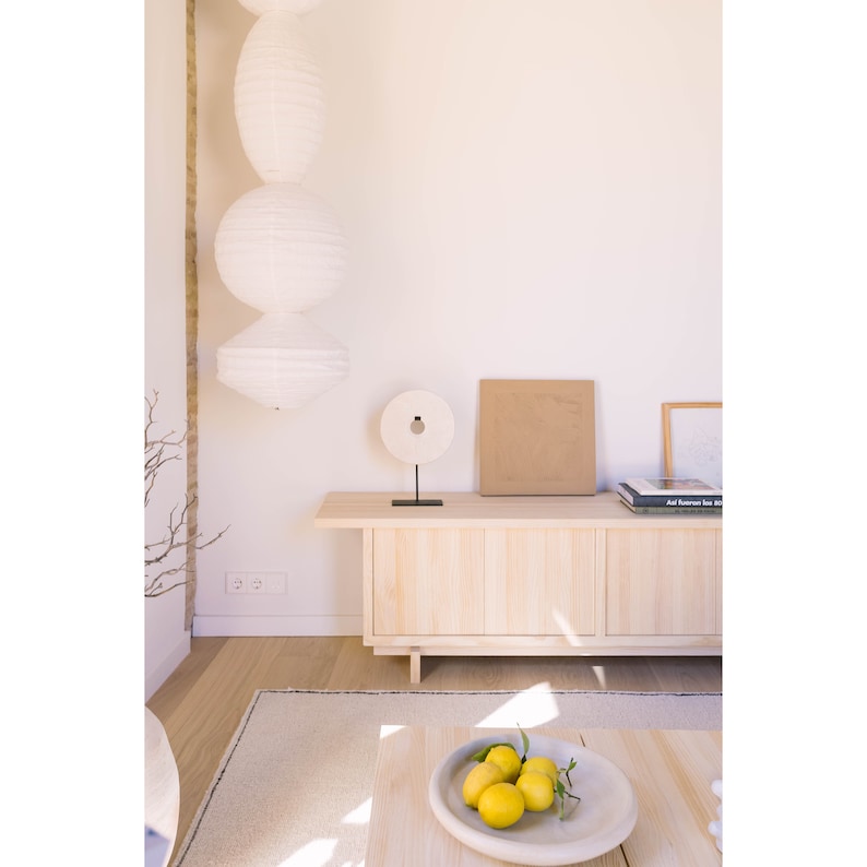 May include: A light wood cabinet with three doors and a white stone circle sculpture on top. The cabinet is against a white wall with a white paper lantern hanging from the ceiling. There are two books and a framed picture on top of the cabinet.