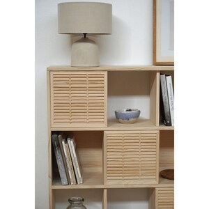 May include: A light wood bookshelf with slatted doors and open shelves. The shelves are filled with books and a small blue bowl. A lamp with a beige shade sits on top of the bookshelf.