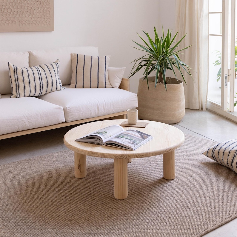 May include: A light wood round coffee table with four cylindrical legs sits on a beige rug. A white sofa with striped pillows is in the background. A large potted plant and an open book are also visible.