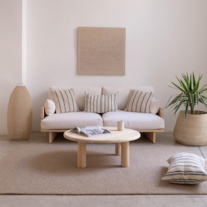 May include: A modern living room setup featuring a light-colored sofa with striped pillows, a round wooden coffee table, and a woven floor lamp. A potted plant and a textured wall art piece complete the minimalist design.